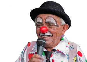 Un clown sorridente con cappello nero, naso rosso e camicia a pois parla in un microfono