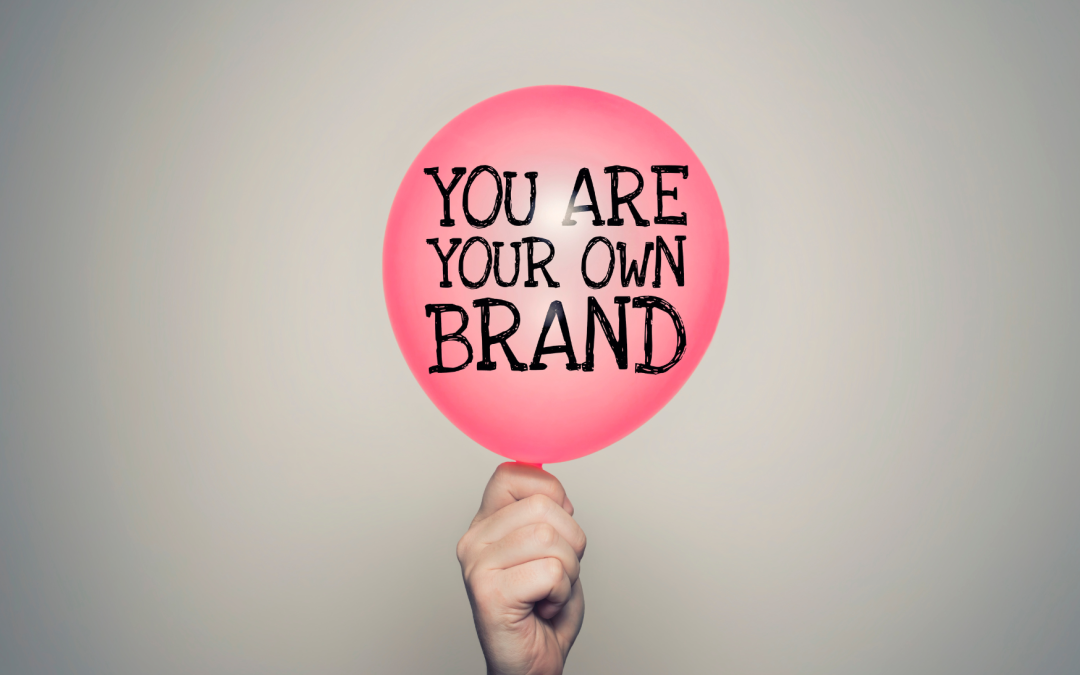 Una mano tiene un palloncino rosa con la scritta “You are your own brand” su sfondo grigio chiaro