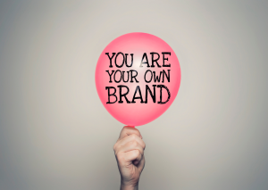 Una mano tiene un palloncino rosa con la scritta “You are your own brand” su sfondo grigio chiaro